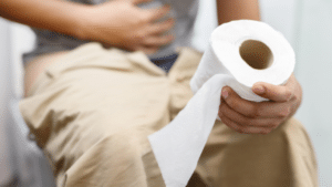 A close-up, cropped shot of a person sitting on a toilet with their khaki pants pulled down, holding a roll of white toilet paper in their right hand while resting their left hand on an aching stomach.