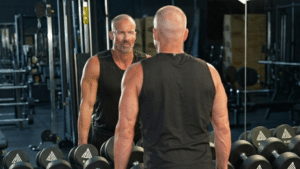 A muscular, bald man wearing a black tank top stands in a gym, looking at his reflection in a large mirror behind a row of dumbbells.