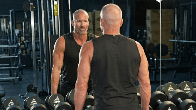 A muscular, bald man wearing a black tank top stands in a gym, looking at his reflection in a large mirror behind a row of dumbbells.
