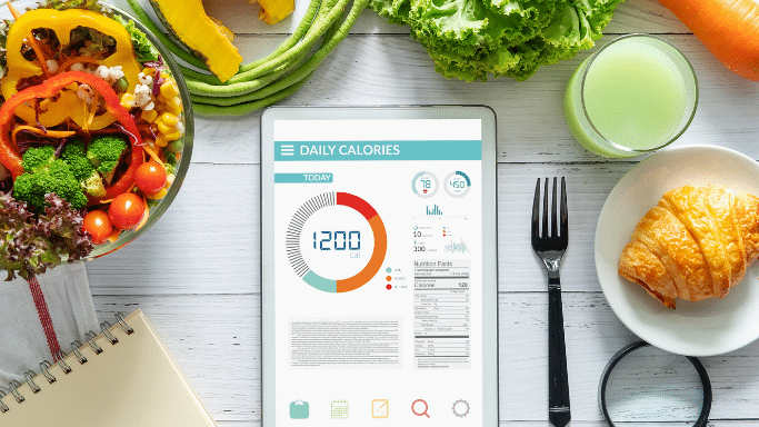 A top-down view of a tablet displaying a daily calorie tracker showing 1,200 calories, surrounded by a salad, a croissant, a green smoothie, and fresh vegetables on a white wooden table.