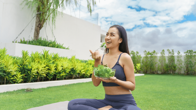 A smiling woman in grey athletic wear sits cross-legged on a lawn, holding a bowl of salad and eating a forkful of greens.