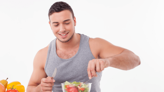 A smiling young man in a grey tank top stands against a white background, using a knife and fork to prepare a fresh vegetable salad.