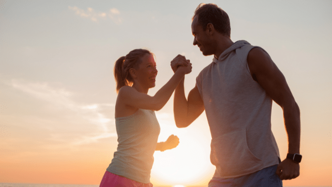 A man and woman in athletic clothing share an enthusiastic high-five or arm lock against a warm, glowing sunset background.