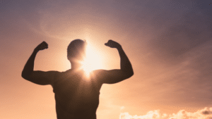 Silhouette of a person from the back, flexing both arms to show off their biceps against a bright sunset. The sun is positioned directly behind the person's head, creating a strong lens flare and sunburst effect. The sky transitions from a deep purple at the top to a warm orange and yellow near the horizon, with light clouds scattered at the bottom right.
