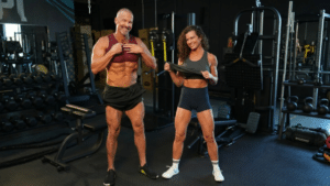 A fit, smiling middle-aged man and woman posing side-by-side in a gym, lifting their shirts to show defined abdominal muscles.