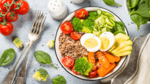 A colorful healthy bowl filled with buckwheat, broccoli, carrots, avocado, cherry tomatoes, sliced hard-boiled eggs, and cucumber, set next to fresh tomatoes and forks.