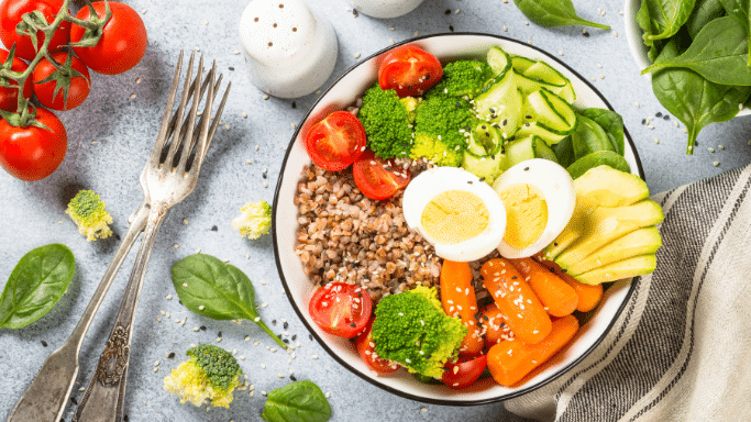 A colorful healthy bowl filled with buckwheat, broccoli, carrots, avocado, cherry tomatoes, sliced hard-boiled eggs, and cucumber, set next to fresh tomatoes and forks.