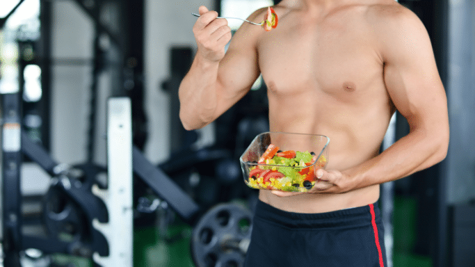 A shirtless, muscular man stands in a gym holding a glass container of fresh salad and lifting a forkful of food toward his mouth.