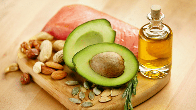 A close-up arrangement of healthy fats on a wooden board, featuring a halved avocado with its pit, a small bottle of golden oil, a piece of raw salmon, and an assortment of nuts and seeds.