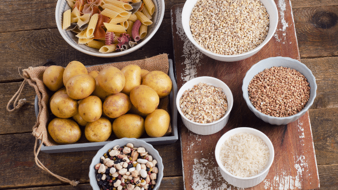 A top-down view of various carbohydrate-rich foods arranged on a rustic wooden surface, including a crate of small yellow potatoes, colorful pasta, rice, oats, buckwheat, and mixed beans.