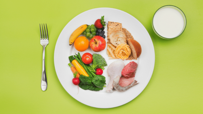 A top-down view of a balanced meal on a white plate divided into sections of fruits, vegetables, grains, and proteins, next to a glass of milk and a fork on a bright green background.