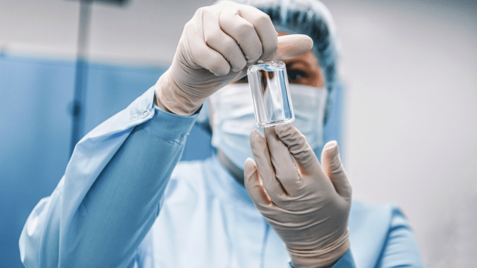 A laboratory technician wearing a mask, cap, and sterile gloves holds up a clear glass vial containing a liquid for inspection.