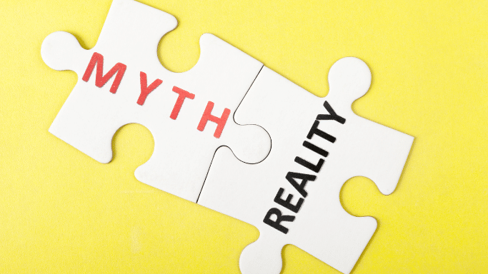 Two white jigsaw puzzle pieces lying on a yellow surface fit together, with the word "MYTH" printed in red on one and "REALITY" in black on the other.