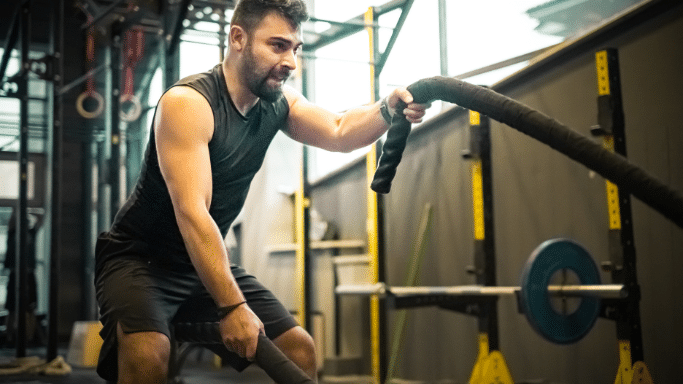 A focused man in athletic wear grips heavy battle ropes during a high-intensity workout inside a gym.