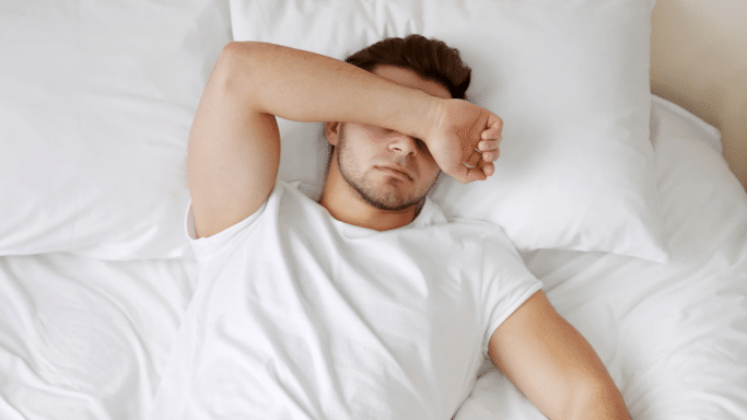 A young man wearing a white t-shirt lies flat on his back in bed, covering his eyes with his forearm.