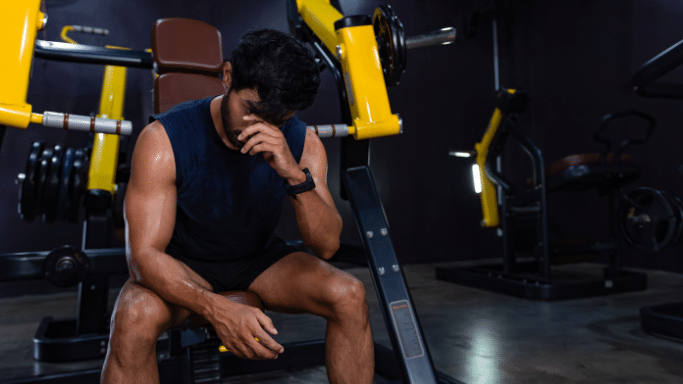 An athletic man sits on a gym machine, looking down with his hand pressed to his forehead in a moment of exhaustion or frustration.