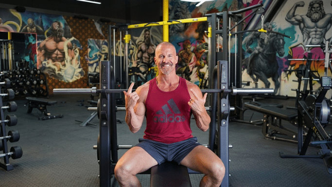 A smiling, muscular man in a red Adidas tank top sits on a weight bench in a gym, gesturing with his hands.