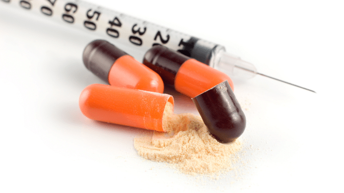 A close-up shot showing several orange and dark brown capsules lying next to a thin medical syringe on a white surface. One capsule is broken open, spilling a pile of light-beige powder onto the surface.