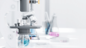 A close-up, shallow-depth-of-field shot of a white and grey compound microscope in a bright laboratory. In the blurred background, various glass beakers and flasks containing blue and pink liquids sit on a white lab bench.