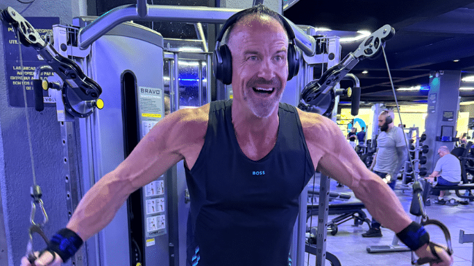 A middle-aged man with a grey goatee and a joyful expression performs a chest fly exercise on a cable machine. He is wearing over-ear headphones, a black tank top, and wrist wraps. The background shows a modern, dimly lit commercial gym with other patrons and equipment.