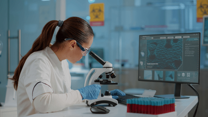 A female scientist with her hair in a ponytail wears a white lab coat, blue nitrile gloves, and safety glasses while working in a high-tech lab. She is looking through a microscope with one hand on a computer keyboard. A monitor next to her displays a magnified cellular image.
