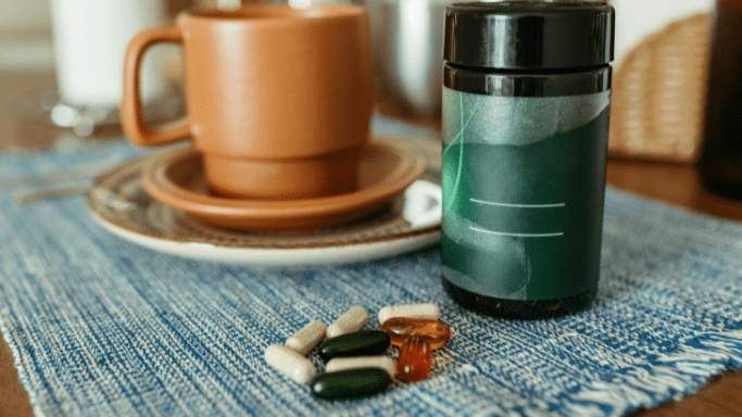 A variety of health supplements, including green capsules and amber softgels, are scattered on a blue woven placemat. In the background, a dark green supplement bottle stands next to a brown ceramic coffee mug and saucer.