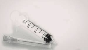 A close-up shot of a clear plastic medical syringe with black volume markings up to 3 ml. A separate, thin hypodermic needle with a clear protective cap lies next to it on a reflective white surface.