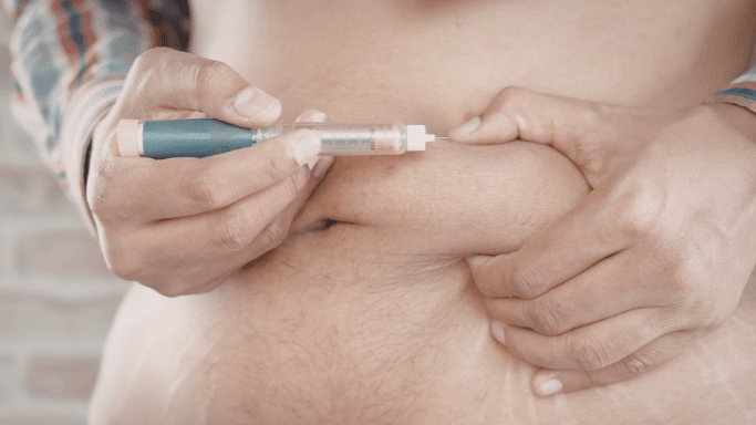 A close-up shot of a person's torso as they perform a subcutaneous injection into their abdomen. One hand pinches a fold of skin while the other hand holds the pen steady against the injection site.