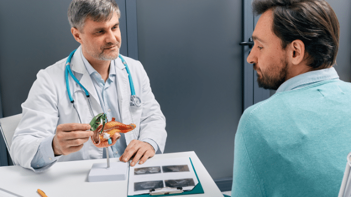 A doctor in a white lab coat and blue stethoscope sits at a desk, explaining a medical model of a pancreas and gallbladder to a male patient. A clipboard with X-ray images sits on the desk between them in a bright office setting.