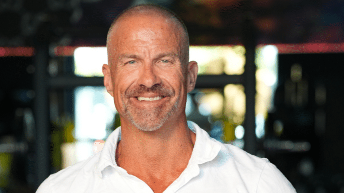 A head-and-shoulders portrait of a middle-aged man with a short buzz cut and a salt-and-pepper goatee. He is wearing a white collared shirt and has a kind, confident smile. The background is a soft-focus indoor environment, likely a gym.