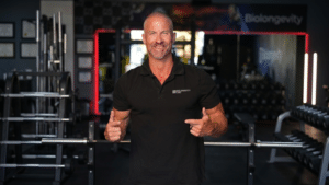 A muscular, smiling man with a shaved head stands in a gym wearing a black polo shirt with "Biolongevity Labs" on the chest. He is pointing both index fingers toward himself in front of a squat rack.