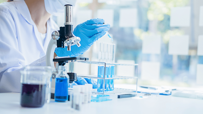 Close-up of a scientist in a white lab coat and blue gloves using a pipette to drop liquid into a test tube. A microscope and various blue-liquid-filled vials are on the table in a bright, modern lab.
