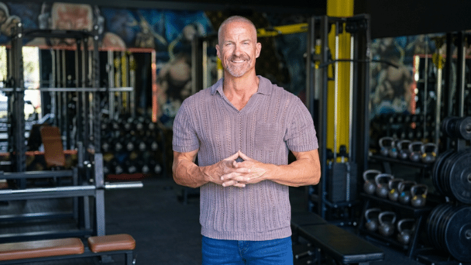 A muscular man with a shaved head and graying beard stands in a gym, smiling with his hands clasped. He is wearing a mauve textured polo shirt and blue jeans, with kettlebells and weight machines visible in the background.