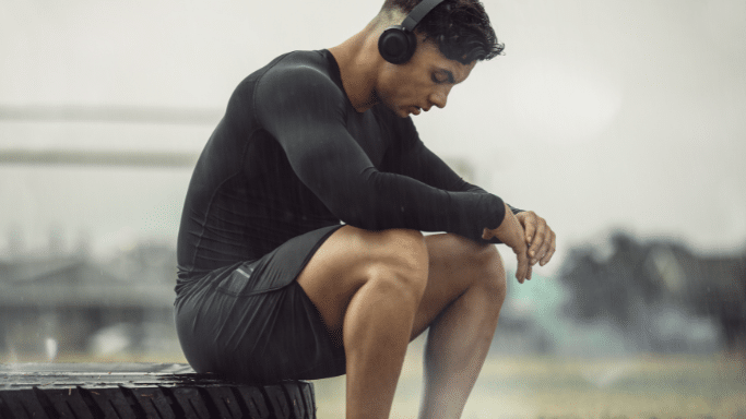A man in a black long-sleeved athletic shirt and shorts sits on a large tire outdoors in the rain. He is wearing headphones and looking down, appearing to rest or focus during a workout.