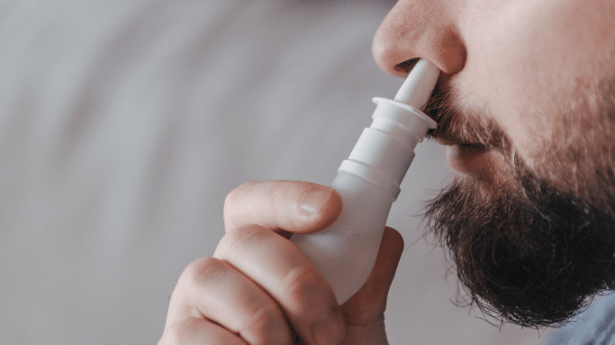 A close-up profile view of a bearded man using a white nasal spray bottle. The nozzle is inserted into one nostril as he prepares to administer the medication.