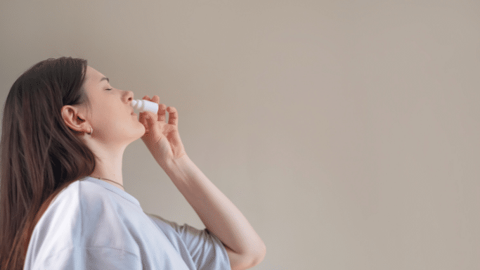 A profile shot of a young woman with dark hair tilting her head back slightly while using a white nasal spray bottle.