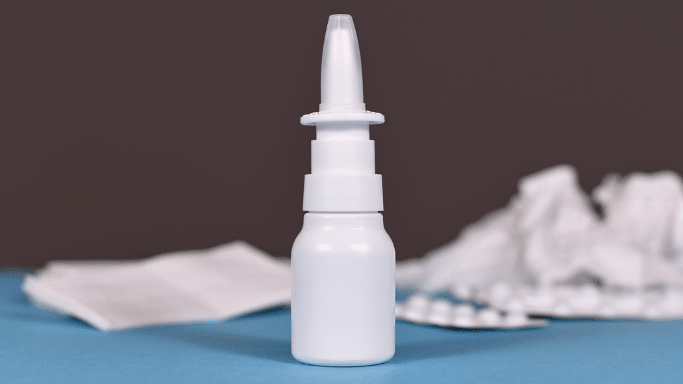 A plain white nasal spray bottle stands centered on a blue surface. In the blurred background, there are crumpled white tissues and a blister pack of pills.