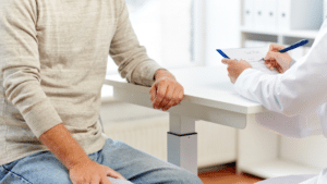 Close-up of a patient in a tan sweater and jeans sitting at a white desk opposite a healthcare provider. The provider, seen only by their arm in a white lab coat, is using a blue pen to write on a paper