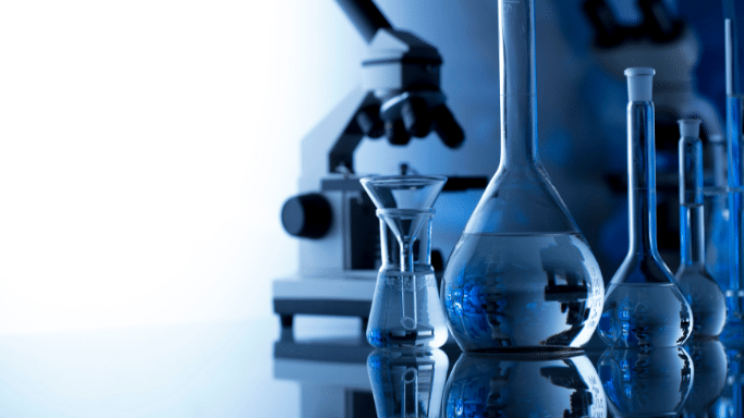 A close-up of a laboratory bench set against a blurred, glowing blue background. In sharp focus are various glass beakers and flasks filled with blue liquid, reflecting on the black tabletop. In the background, a large microscope is slightly out of focus.