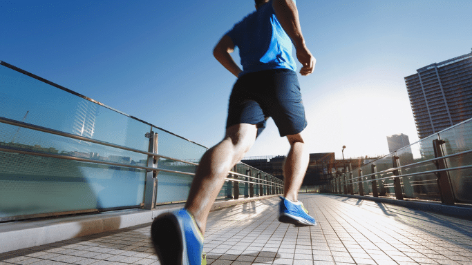 A low-angle, back view of a man in a blue t-shirt and dark athletic shorts. He is running on a modern pedestrian bridge with glass railings. The image is a action shot with motion blur, and a city building is visible in the distance under a bright blue sky.