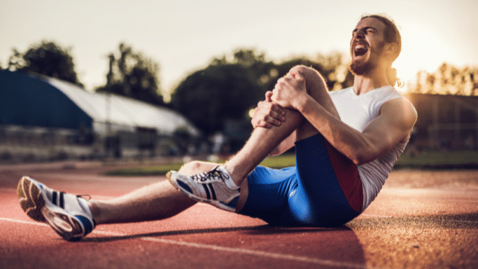 A long-haired man with a beard, wearing a white t-shirt and blue athletic shorts, is sitting on a red running track at sunset. He is holding his right knee with both hands, his mouth open in a grimace of pain.