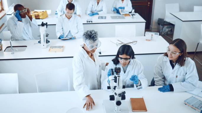 An elevated view of a busy science lab with white counters. In the foreground, three scientists in lab coats examine a microscope. In the background, other scientists are busy writing, looking at a laptop, and working.