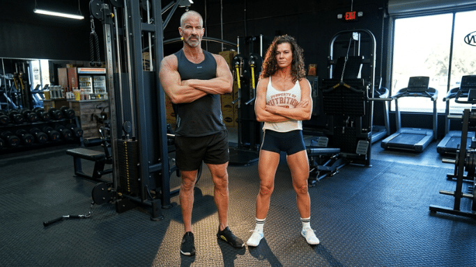 A full-body shot of a muscular man and woman standing confidently in a brightly lit gym. The man, on the left, has short grey hair and is wearing a black tank top and shorts with his arms crossed. The woman, on the right, has long curly hair and is wearing a white graphic tank top and dark shorts, also with her arms crossed. Various gym equipment, including a cable machine and treadmills, are visible in the background.