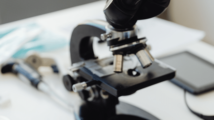 A close-up, slightly blurred photograph of a black laboratory microscope. The focus is on the objective lenses and the stage, while the background—containing various lab tools and a tablet—is soft and out of focus, creating a professional scientific atmosphere.