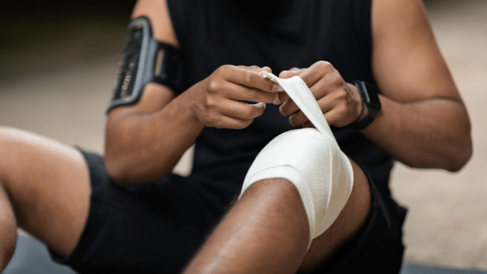 A close-up shot of a person in athletic wear sitting down and carefully wrapping a white compression bandage around their knee. They are wearing a fitness tracker on their wrist and an arm-mounted phone carrier, suggesting a sports-related injury or preventative care during a workout.