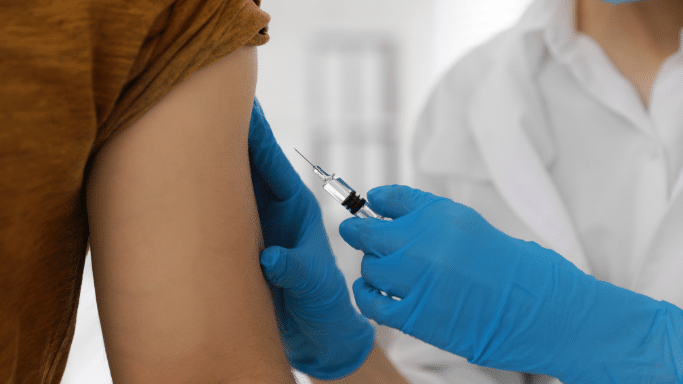 A close-up shot showing a healthcare worker in blue nitrile gloves and a white coat about to administer an injection into a patient's upper arm. The patient is wearing a textured brown shirt.