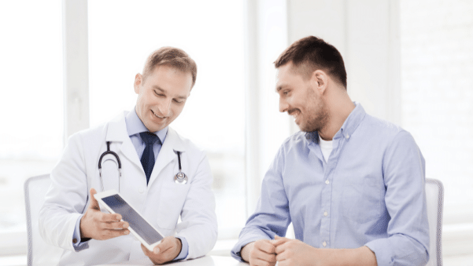 A male doctor in a white lab coat and stethoscope smiles as he shows a digital tablet to a male patient. Both are seated at a white table in a bright, modern office.