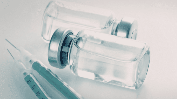 A still life of two glass medical vials filled with clear liquid resting next to two syringes with green plungers. The image has a cool, clinical blue-grey tint.