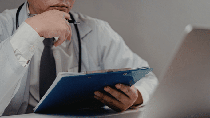 A close-up of a doctor in a white coat and tie, sitting at a desk and holding a blue clipboard. His hand is on his chin in a pensive gesture as he looks toward a laptop screen.
