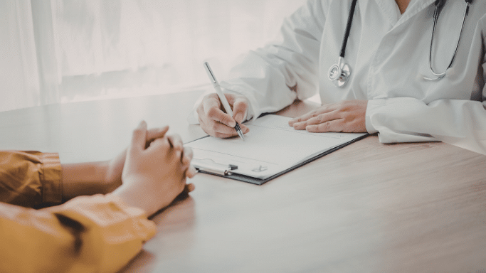 A doctor's hands are shown writing on a medical form on a clipboard. Across the desk, a patient's hands are clasped together. Only the arms and torsos of both individuals are visible.